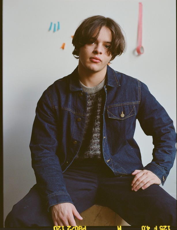 Young adult in dark denim jacket and trousers sits indoors against a white wall with art; soft lighting and relaxed pose.