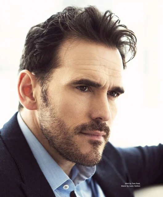 Portrait of a dark-haired man with a beard, wearing a blue collared shirt and a dark blazer, looking off to the side with furrowed brows.