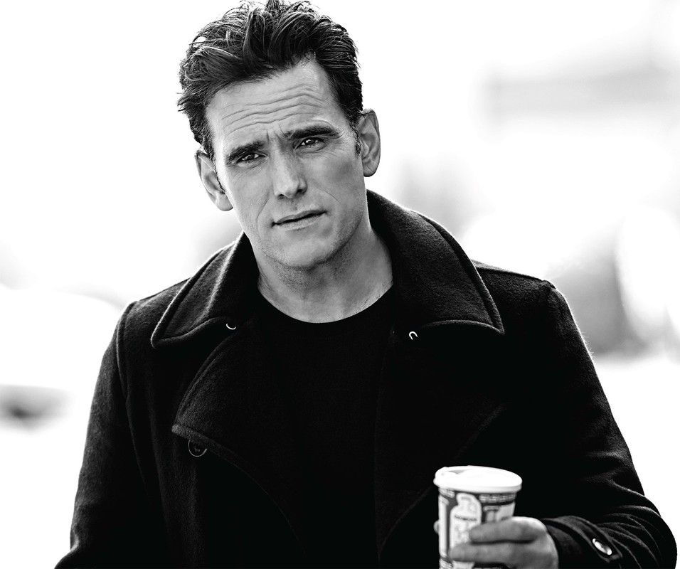 Portrait of an adult male in a black coat holding a coffee cup, photographed outdoors in black and white.