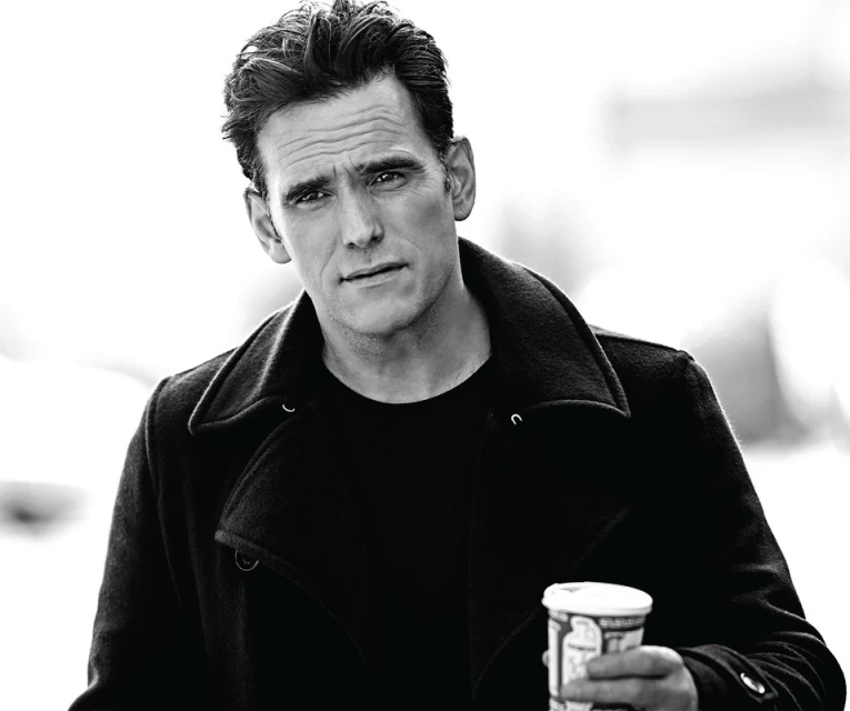 Portrait of an adult male in a black coat holding a coffee cup, photographed outdoors in black and white.