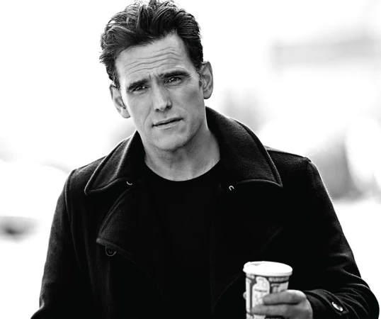 Portrait of an adult male in a black coat holding a coffee cup, photographed outdoors in black and white.