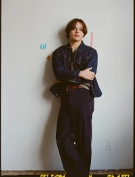 Young man in a denim jacket and dark trousers leaning against a white wall with colorful paper tabs