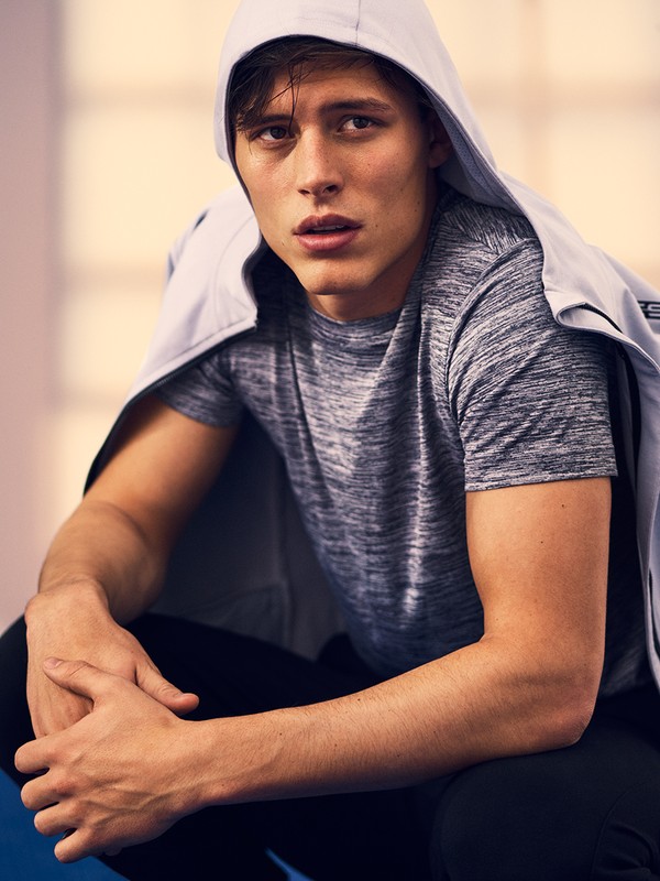Portrait of a young adult male crouching indoors, wearing a gray t-shirt and a light gray hooded jacket, gazing forward with a neutral expression.