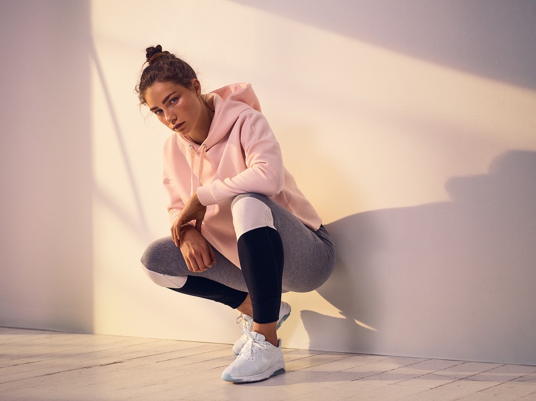 Young woman wearing a pink hoodie and athletic leggings crouching in a studio with warm light.