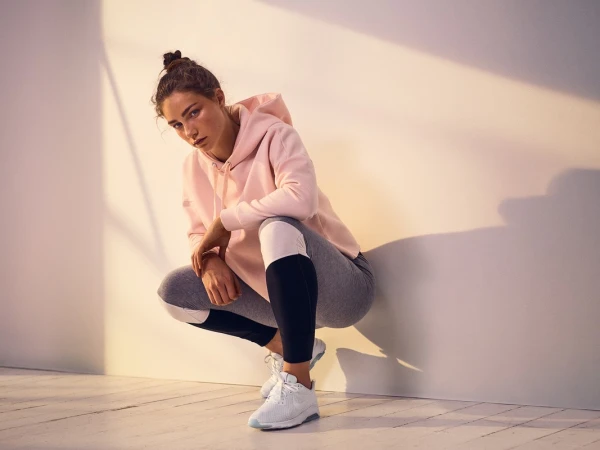Young woman wearing a pink hoodie and athletic leggings crouching in a studio with warm light.