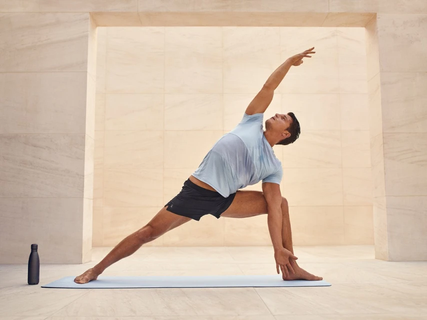 A man on a blue yoga mat is doing an extended triangle yoga pose in a bright, minimalist, indoor studio with beige walls.