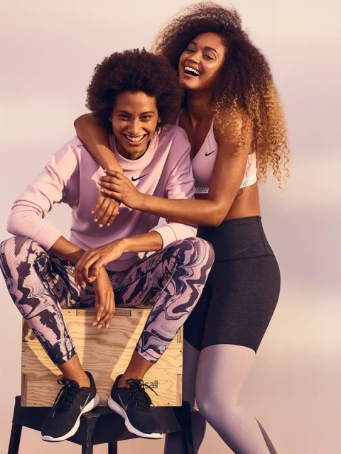 Two women smile in workout gear. One sits on a wooden crate wearing lilac sweatshirt and patterned leggings. The other woman stands beside her.