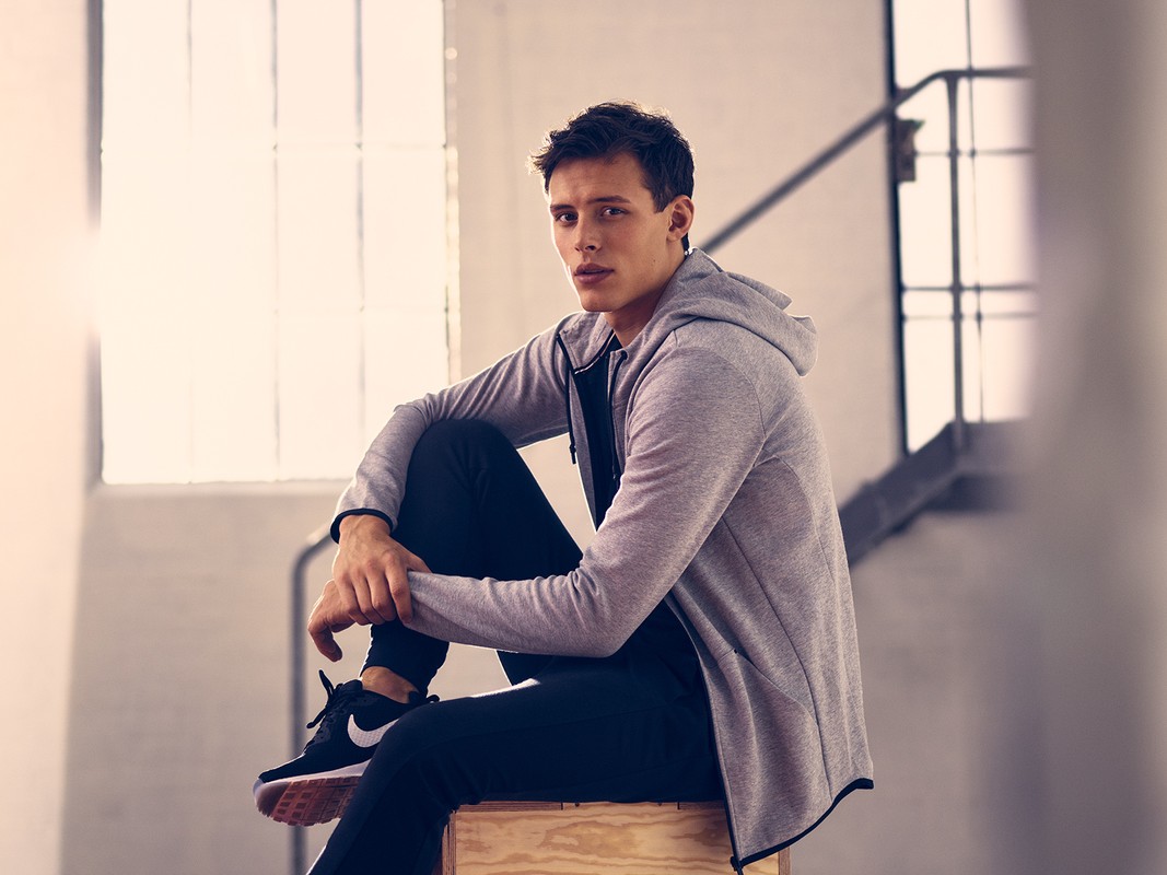 A young adult male sits in a grey hoodie and black athletic pants on a wooden block indoors with natural light from a nearby window.