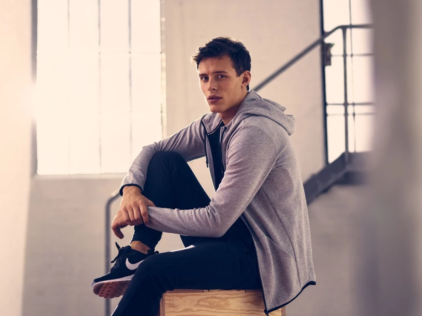 A young adult male sits in a grey hoodie and black athletic pants on a wooden block indoors with natural light from a nearby window.