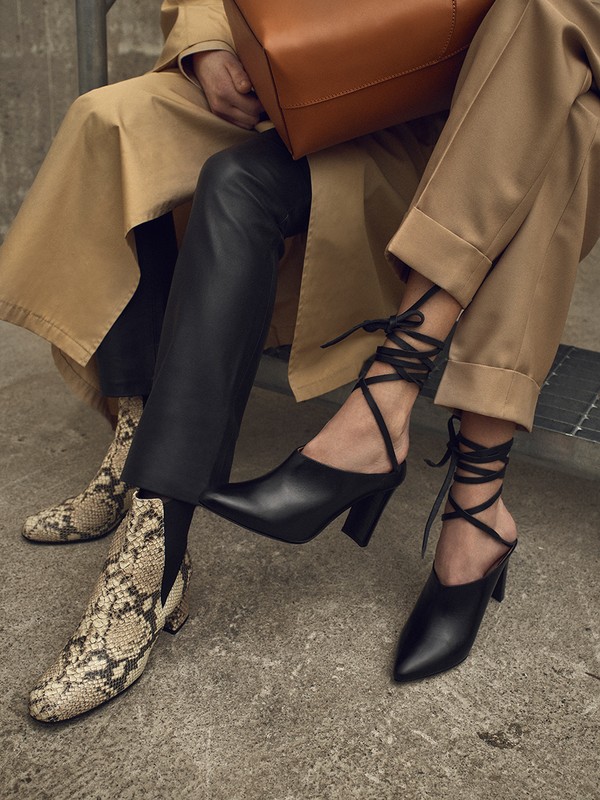 Fashion image showcasing a woman wearing black leather pants, a tan trench coat, and black high heels with a ribbon design. 