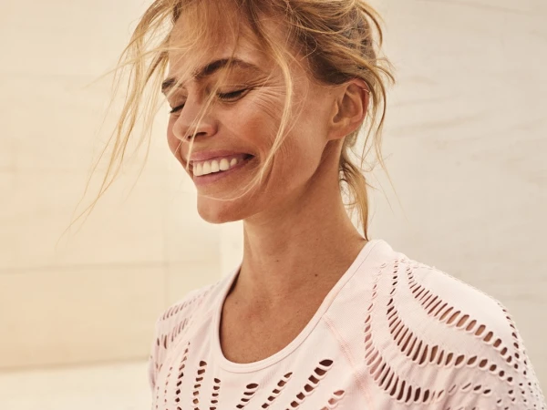 Close-up of a smiling adult woman with blond hair wearing a light pink shirt; portrait oriented, shot indoors, soft lighting.