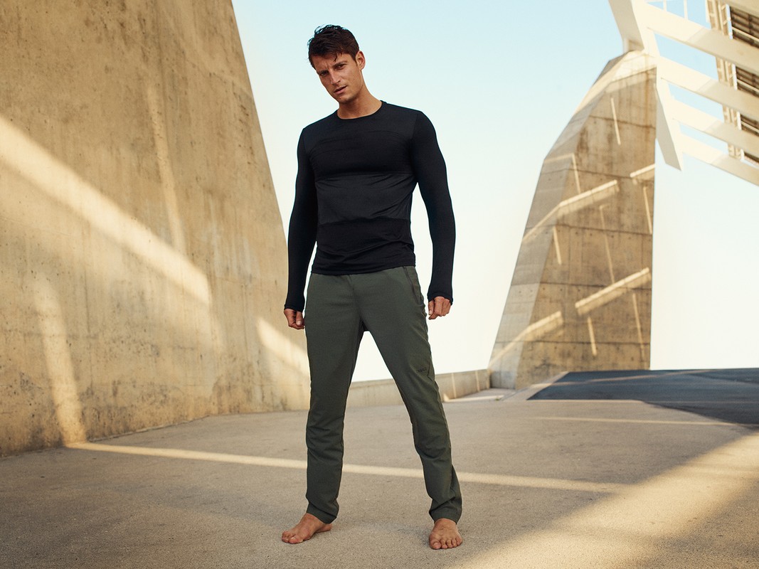 Athletic man in long-sleeved shirt and sweatpants stands barefoot in an urban setting with a modern concrete structure in the background.