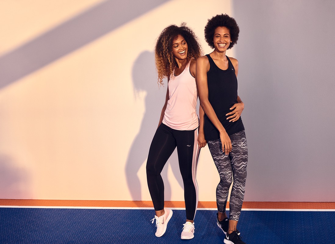 Two women in athletic wear smile indoors against a two-tone wall and floor. One wears a peach tank and the other wears a black tank.