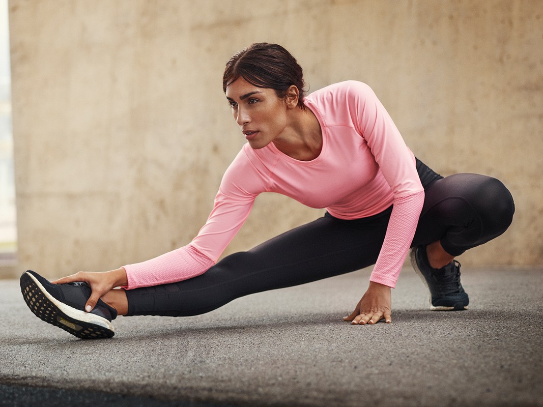 Athletic woman in pink top and black leggings stretches her leg, touching her toes, in a fitness or training studio setting.