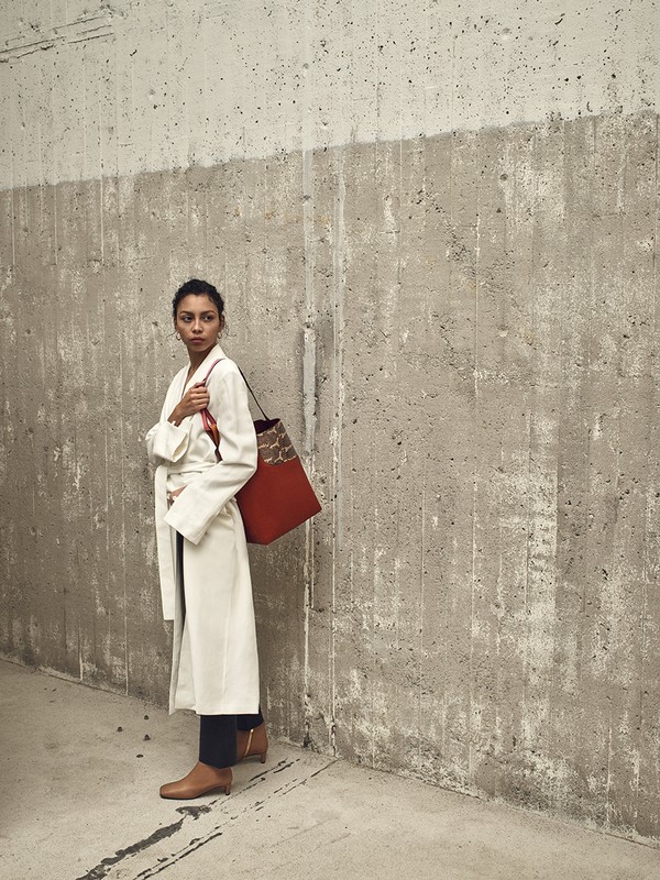 A woman in a cream coat stands against a concrete wall carrying a red tote bag, showcasing a blend of fashion and urban textures.