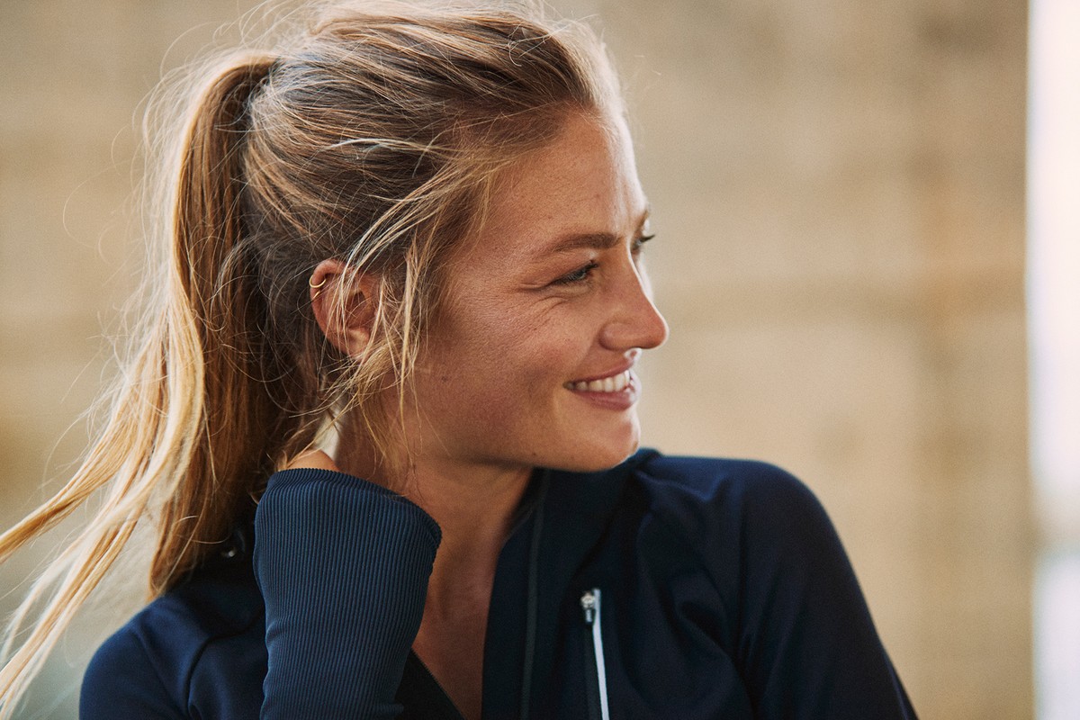 Close-up of a blonde woman with a ponytail wearing a blue athletic jacket looking to the side in a brightly lit, modern indoor setting.