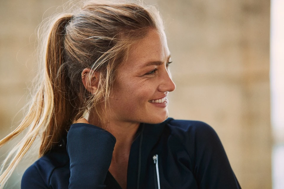 Close-up of a blonde woman with a ponytail wearing a blue athletic jacket looking to the side in a brightly lit, modern indoor setting.