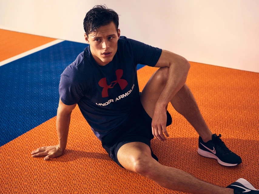 Adult male athlete in sportswear sits on a textured orange and blue floor in an indoor setting, wearing an Under Armour shirt and athletic shoes.