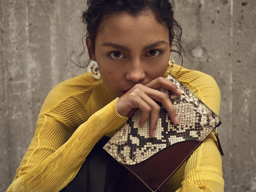 Woman in yellow poses indoors with snakeskin clutch and stylish earrings against a concrete wall in a medium indoor product shot.