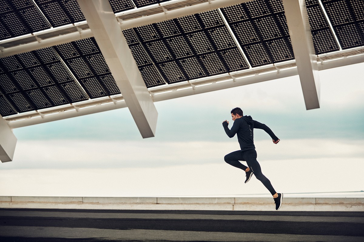 A man in black athletic wear sprints mid-air in an outdoor setting with a modern architectural structure overhead, under a bright sky.