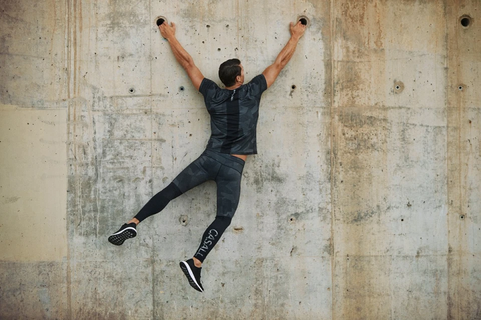 Athletic man hanging on a concrete wall in sportswear. He's wearing a dark camo shirt and leggings and performing a strength exercise.