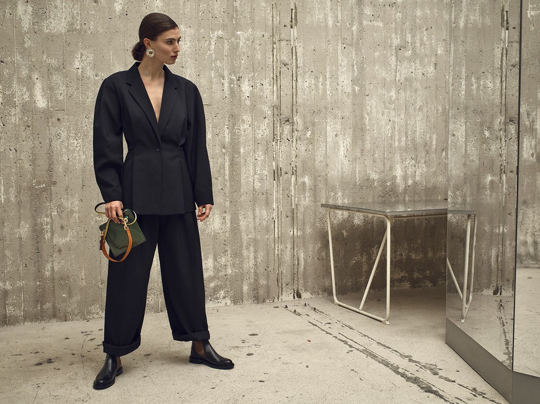 Adult woman in black pantsuit with olive purse poses in studio against concrete wall with white table and mirror, fashion portrait.