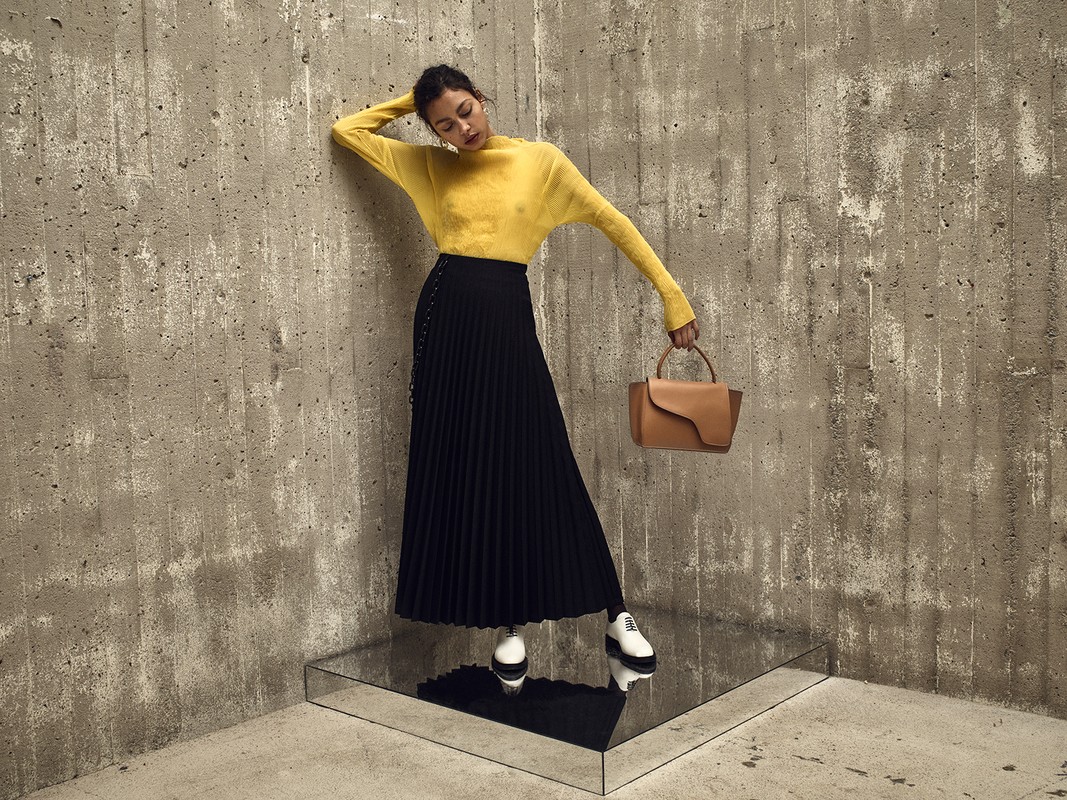 Woman in yellow sweater and black skirt poses with a handbag in front of a textured concrete wall on a reflective platform.