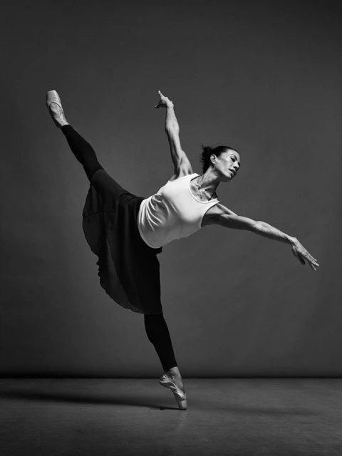 Monochrome photograph featuring an adult female ballet dancer in an arabesque pose, wearing pointe shoes, a tank top, and a skirt.