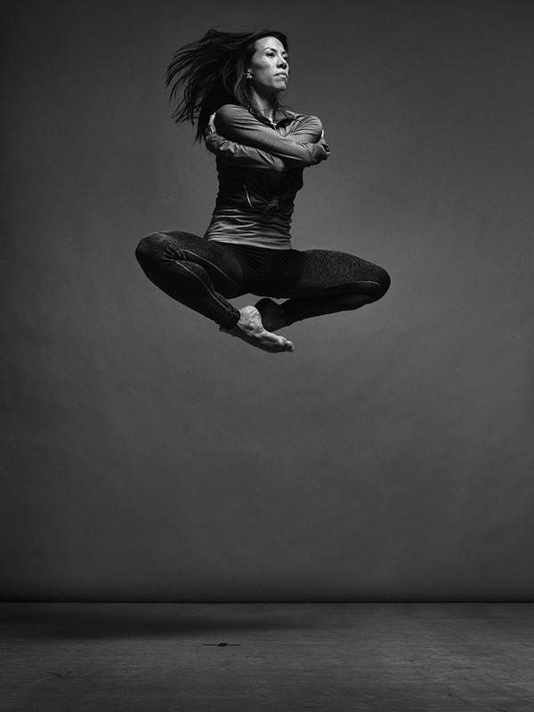 A woman in athletic wear leaps against a grey backdrop, her arms crossed and hair flying, captured in a black and white photo studio shot.