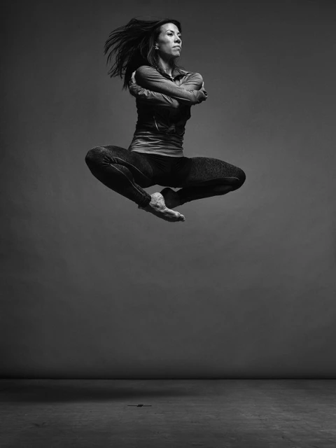 A woman in athletic wear leaps against a grey backdrop, her arms crossed and hair flying, captured in a black and white photo studio shot.