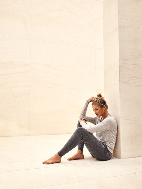 Young woman sits barefoot on the floor in grey loungewear with her head resting on her arm in an indoor minimalist setting.