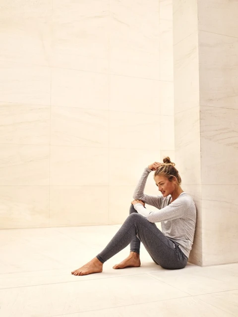 Young woman sits barefoot on the floor in grey loungewear with her head resting on her arm in an indoor minimalist setting.