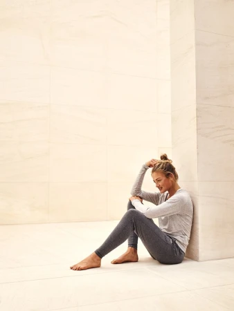 Young woman sits barefoot on the floor in grey loungewear with her head resting on her arm in an indoor minimalist setting.