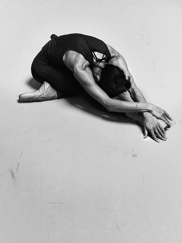 Black-and-white studio shot of a single athletic adult in a tank top, pointe shoes and dark pants, seated and stretching forward.