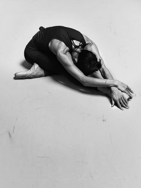 Black-and-white studio shot of a single athletic adult in a tank top, pointe shoes and dark pants, seated and stretching forward.