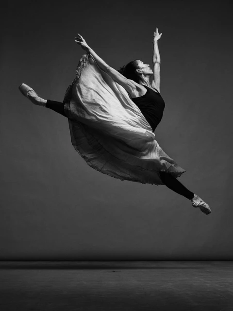 Monochrome studio shot of a ballet dancer leaping in the air, wearing pointe shoes, a black tank top, and a long, flowing skirt.