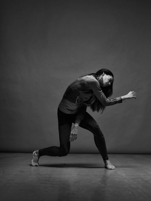 Full-length portrait of a barefoot female dancer with long dark hair, dressed in athletic attire, captured in a dramatic pose in the studio.