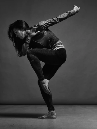 Monochrome studio shot of a woman in athletic wear striking a dynamic pose, arms extended, one leg raised, showcasing a blend of strength and flexibility.
