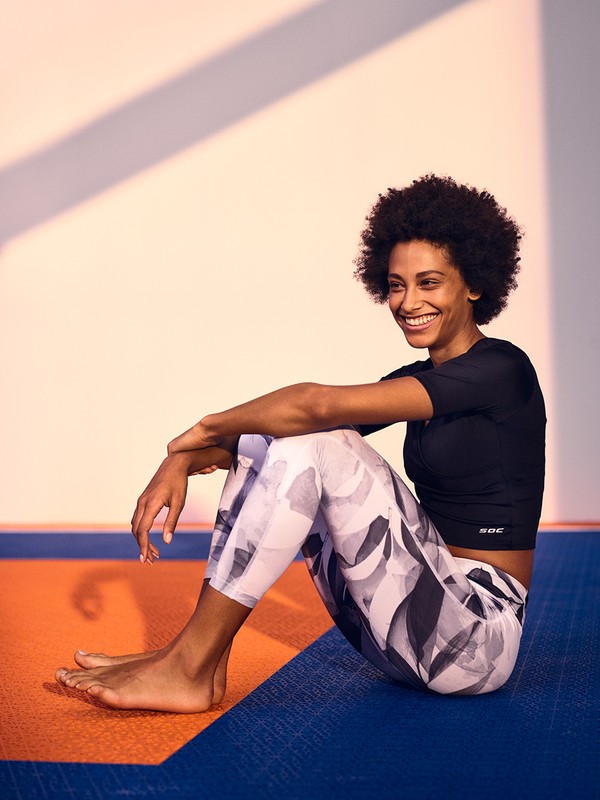 Smiling woman with afro sits cross-legged on a mat in a black top and printed leggings, enjoying light and promoting active lifestyle.