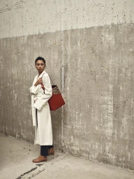 Woman in white belted coat and dark trousers carrying a large red tote bag against a raw concrete wall.