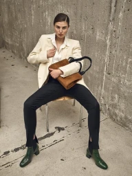 Woman seated on a chair wearing a cream blazer, white shirt, black trousers, and green boots, holding a brown suede bag.