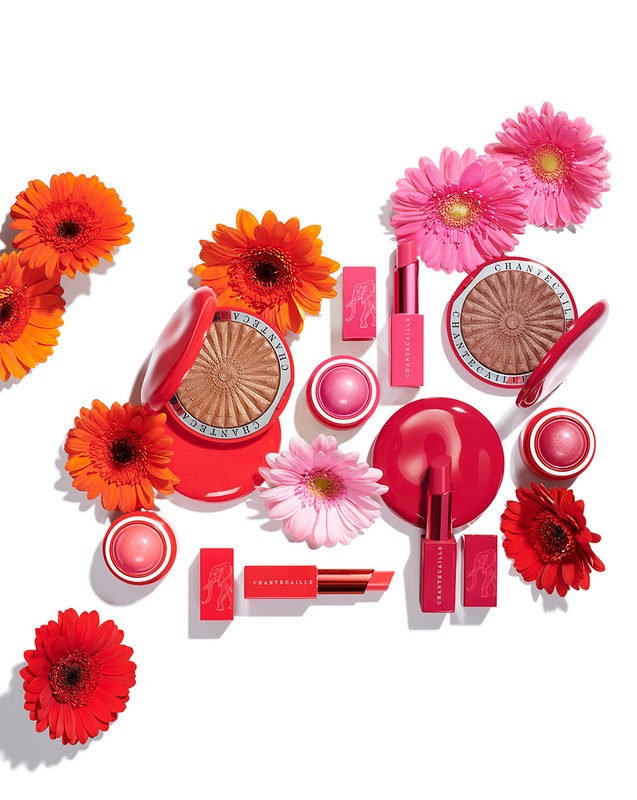 Cosmetic products from Chantecaille arranged with colorful daisy flowers, featuring lipstick, blush, and powder in pink and orange hues.