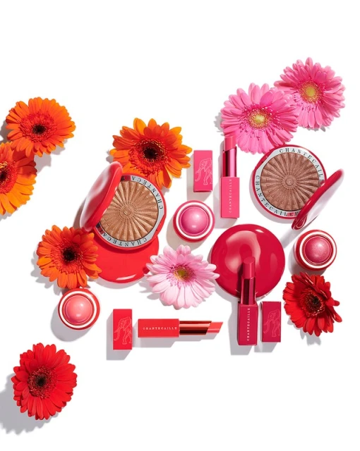 Cosmetic products from Chantecaille arranged with colorful daisy flowers, featuring lipstick, blush, and powder in pink and orange hues.