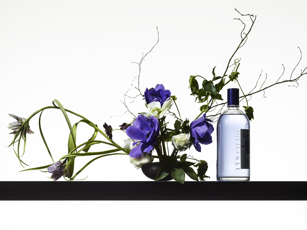 Still life shot of a fragrance bottle with a dark blue cap next to a floral arrangement of purple and white flowers on a black surface.