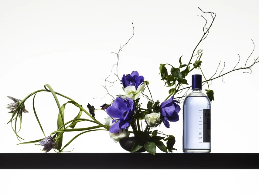 Still life shot of a fragrance bottle with a dark blue cap next to a floral arrangement of purple and white flowers on a black surface.