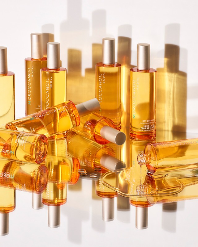 Moroccanoil body oils displayed on a reflective surface, featuring multiple amber-colored glass bottles with silver caps and crisp white background.