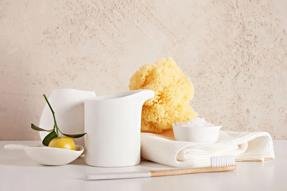 Still life with white porcelain vessels, sea sponge, tangerine, and bamboo toothbrush on a white surface against a beige background.