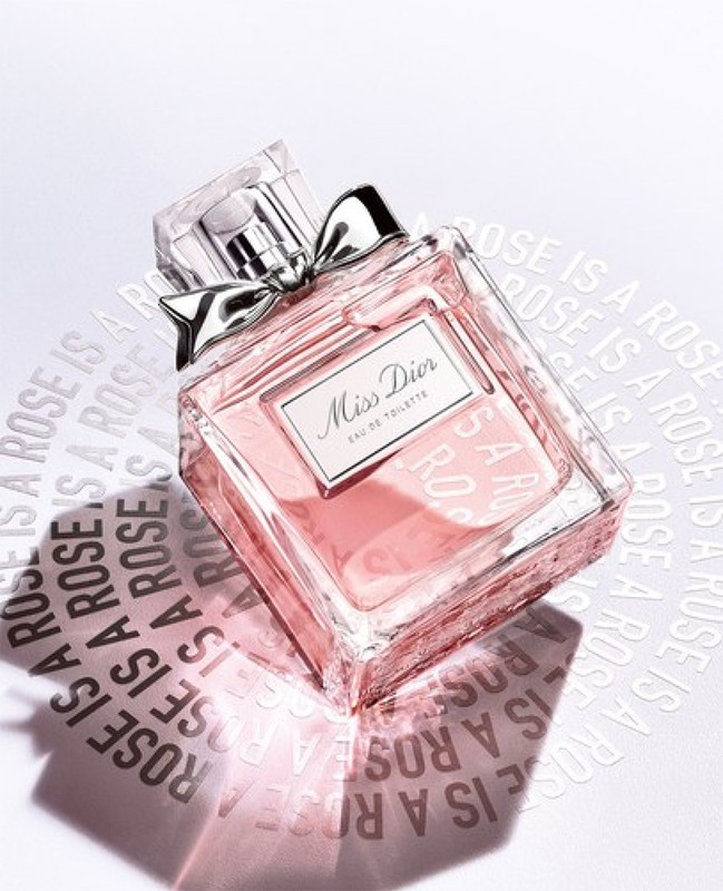 Elevated view of a 'Miss Dior' Eau de Toilette perfume bottle on a white surface with embossed brand messaging, soft light, and a silver bow.