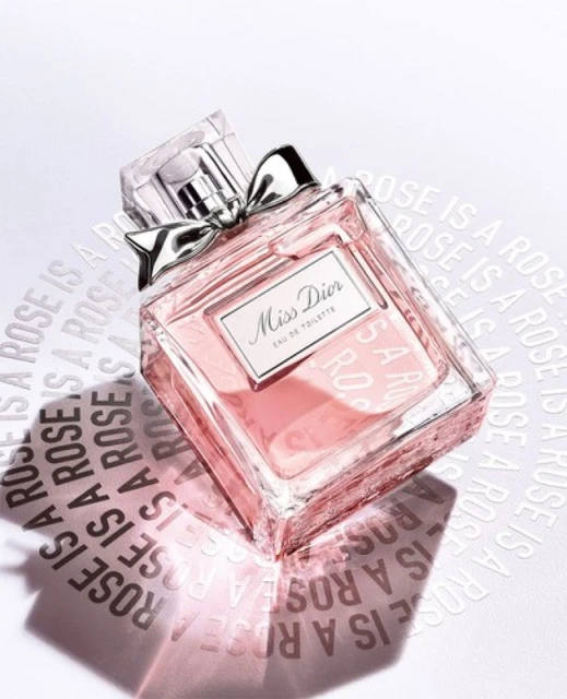 Elevated view of a 'Miss Dior' Eau de Toilette perfume bottle on a white surface with embossed brand messaging, soft light, and a silver bow.