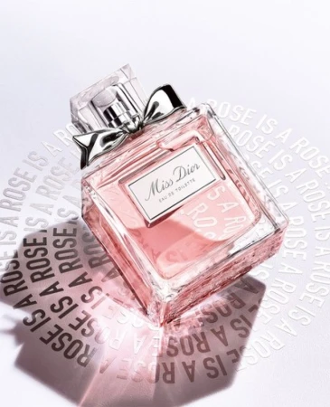 Elevated view of a 'Miss Dior' Eau de Toilette perfume bottle on a white surface with embossed brand messaging, soft light, and a silver bow.